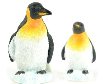 May include: Two penguin figurines, one larger than the other, both with black heads, yellow beaks, orange chests, and white bodies. They are standing on a white base that resembles ice.