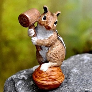May include: A small, brown and white figurine of a chipmunk holding a wooden mallet. The chipmunk is standing on a brown, round, walnut-shaped base.