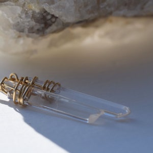 May include: A gold wire wrapped clear quartz crystal pendant necklace. The pendant is made of two quartz crystals that are wrapped together with gold wire. The necklace is on a white surface.