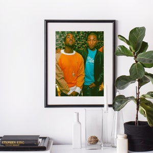 Puede incluir: Una fotografía en blanco y negro enmarcada de dos hombres de pie juntos. El hombre de la izquierda lleva un suéter naranja y marrón y una cadena de oro. El hombre de la derecha lleva una camisa azul y una chaqueta negra. El fondo es un patrón de camuflaje.