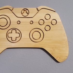 Puede incluir: Una decoración de pared de madera en forma de mando de Xbox, hecha de madera de bambú marrón claro. El mando está grabado con los botones y el logotipo.