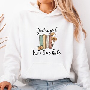 May include: White hoodie with a front pocket and the text "Just a girl Who loves books." The design features a stack of books in green, tan, and pink, with yellow flowers.