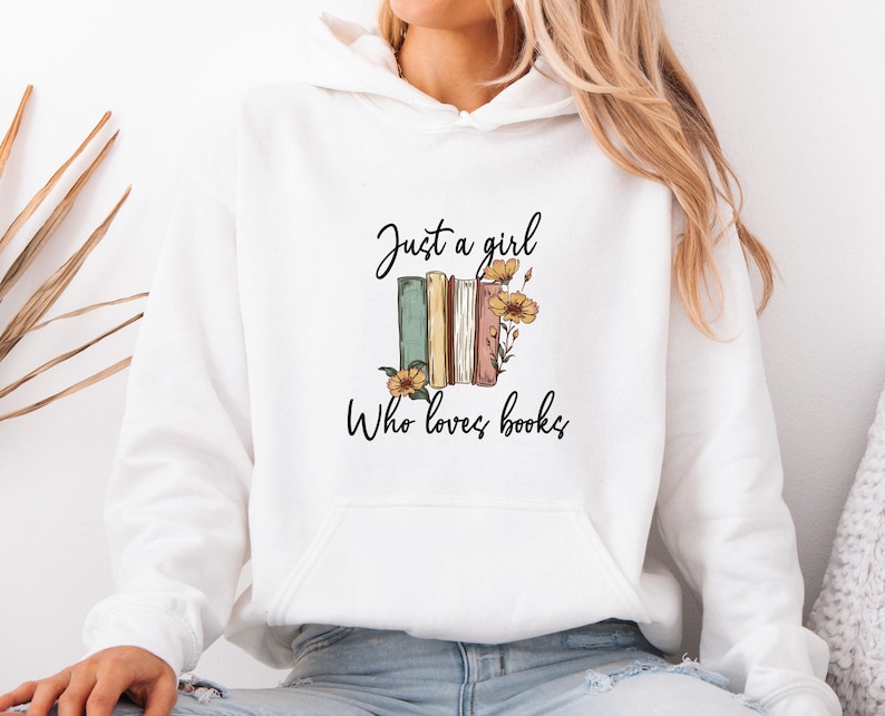 May include: White hoodie with a front pocket and the text "Just a girl Who loves books." The design features a stack of books in green, tan, and pink, with yellow flowers.