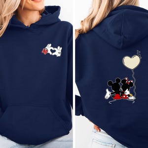 Peut inclure: Sweat à capuche bleu marine avec une poche avant et une capuche. L'avant présente un motif de lapin blanc avec un nœud rouge et un cœur. Le dos affiche un motif Mickey et Minnie avec un ballon en forme de cœur.