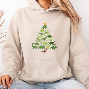 Könnte beinhalten: Beiger Kapuzenpullover mit einem Weihnachtsbaum-Design aus grünen Dinosauriern. Der Baum ist mit Lichterketten geschmückt und mit einem gelben Stern gekrönt. Am Fuß des Baumes befindet sich ein brauner Dinosaurier.
