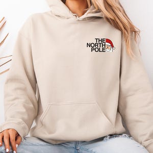 Könnte beinhalten: Beiger Kapuzenpullover mit Fronttasche und Kapuze. Auf der linken Brust befindet sich der Schriftzug "THE NORTH POLE" über einer Weihnachtsmann-Grafik mit roter Mütze. Der Hoodie ist aus weichem Material.
