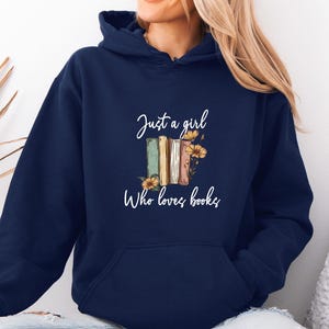 May include: Navy blue hoodie with the text "Just a girl who loves books." The graphic features a stack of books in shades of green, beige, and brown, adorned with yellow flowers. The hoodie has a drawstring hood and a front pocket.