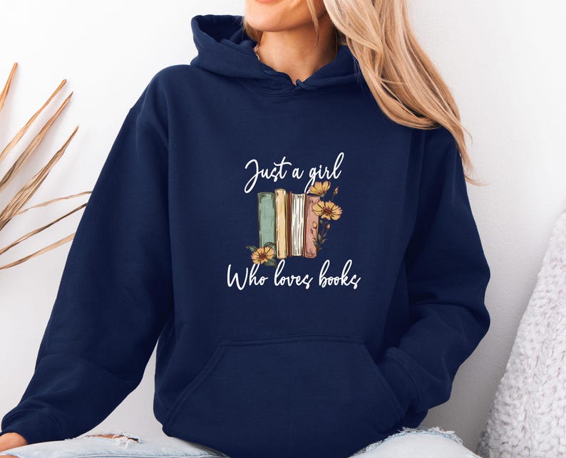 May include: Navy blue hoodie with the text "Just a girl who loves books." The graphic features a stack of books in shades of green, beige, and brown, adorned with yellow flowers. The hoodie has a drawstring hood and a front pocket.