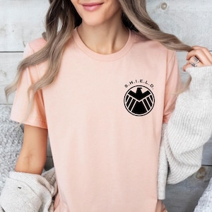 May include: A pink t-shirt with a black S.H.I.E.L.D. logo featuring a falcon and the letters S.H.I.E.L.D.