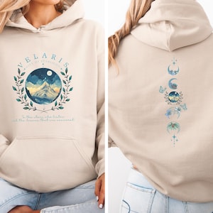 Könnte beinhalten: Beigefarbener Kapuzenpullover mit einem Front-Grafikmotiv einer Berglandschaft in einem kreisförmigen Kranz, dem Text "VELARIS" und einem Zitat. Die Rückseite zeigt ein Design von Mondphasen und Schmetterlingen. Ein gemütliches, lässiges Kleidungsstück.
