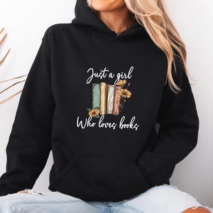 May include: Black hooded sweatshirt with a front pocket and the text "Just a girl Who loves books" in white script. The graphic features a stack of books in various colors with sunflowers.