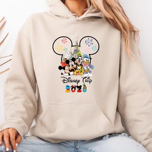Op de afbeelding: Een beige hoodie met een kleurrijke afbeelding van Disney-personages, waaronder Mickey Mouse, Minnie Mouse, Donald Duck en Goofy. Het ontwerp bevat de tekst "Disney Trip" en het jaar "2023". De hoodie heeft een voorzak en een capuchon met trekkoord.