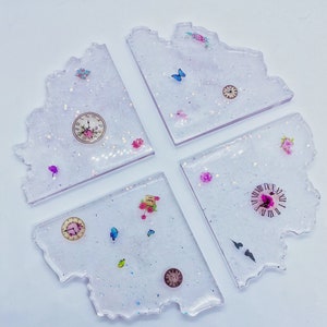 Könnte beinhalten: Set aus vier weißen Harz-Untersetzern mit glitzernder Oberfläche. Jeder Untersetzer hat ein anderes Design mit Uhren, Schmetterlingen und Blumen.