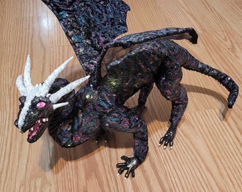 Puede incluir: Un juguete de peluche de dragón negro e iridiscente con cuernos blancos y ojos rosas. El dragón tiene una cola larga y alas.
