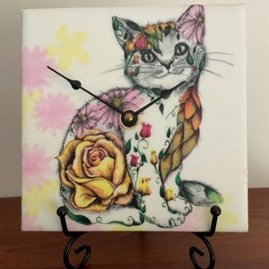 May include: A square clock featuring a whimsical cat design adorned with floral patterns in pink, yellow, and orange. The clock hands are black, and the clock is set on a black metal stand. The cat's fur is white and gray.