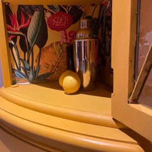 Pode incluir: Um arm&aacute;rio de canto amarelo com uma porta de vidro e uma prateleira forrada com papel de parede de padr&atilde;o tropical. A prateleira cont&eacute;m um coqueteleira de prata e dois lim&otilde;es.