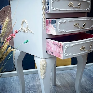 May include: A white painted wooden bedside table with three drawers. The drawers have ornate gold trim and are decorated with vintage paper. The table has four legs with ornate gold trim.