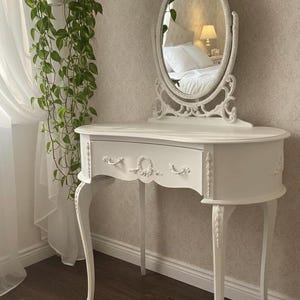 Può includere: Toeletta bianca con specchio ovale e intagli decorativi. Il tavolo ha un fronte curvo con un cassetto singolo e gambe sottili e curve. Una pianta sospesa con foglie verdi è visibile a sinistra.