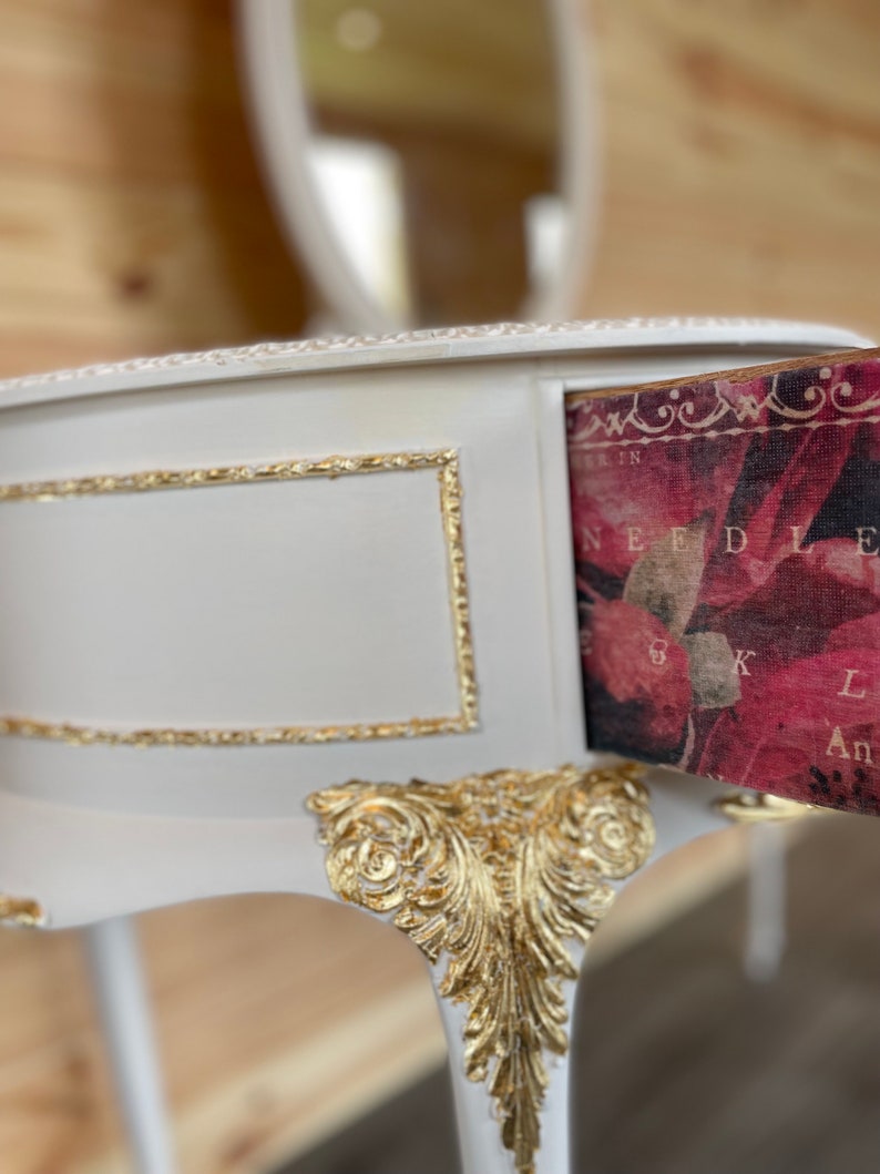 Puede incluir: Una mesa de tocador blanca con detalles dorados. El caj&oacute;n abierto revela un interior con estampado floral y texto. La mesa presenta adornos dorados en las patas y el marco. Un espejo a juego es visible en el fondo.