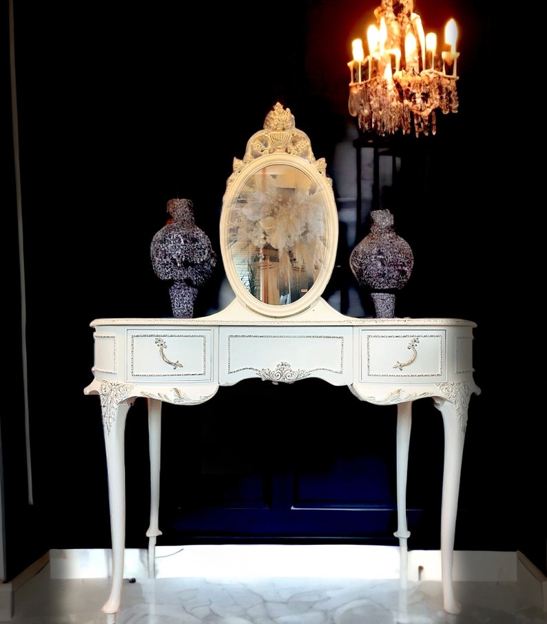 Puede incluir: Una mesa de tocador blanca con un espejo ovalado y dos jarrones decorativos. La mesa tiene tallas ornamentadas y tiradores de cajones. Una l&aacute;mpara de ara&ntilde;a de cristal cuelga sobre la mesa, con un fondo negro.