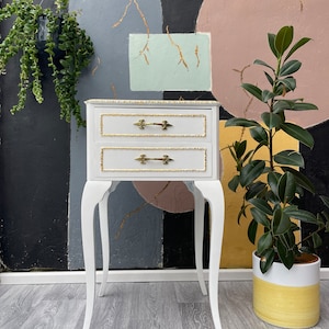 May include: White two-drawer nightstand with gold trim and ornate legs. The nightstand is in front of a wall with abstract shapes painted in pink, blue, and green.