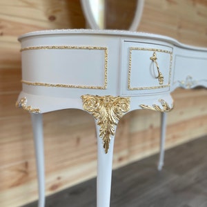 Puede incluir: Una mesa de tocador blanca con detalles dorados. La mesa tiene un caj&oacute;n con un tirador dorado, detalles ornamentales de hojas doradas y un espejo ovalado a juego. La mesa tiene patas curvas y un borde decorativo.