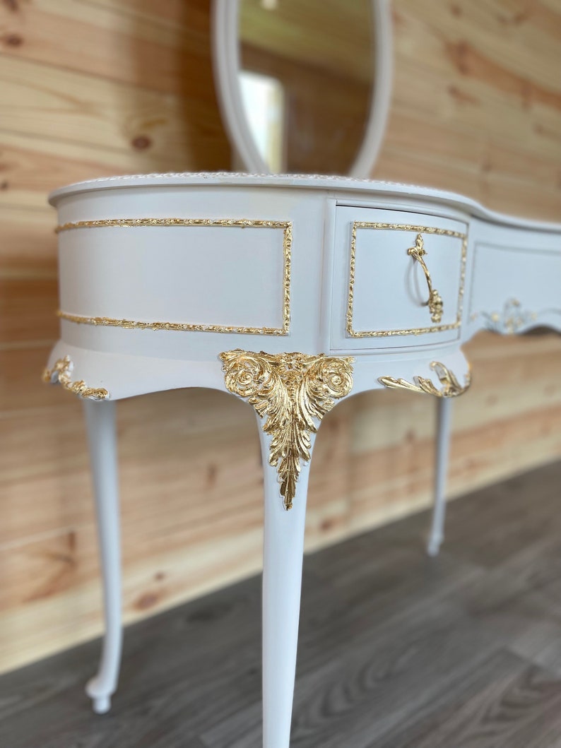 Puede incluir: Una mesa de tocador blanca con detalles dorados. La mesa tiene un caj&oacute;n con un tirador dorado, detalles ornamentales de hojas doradas y un espejo ovalado a juego. La mesa tiene patas curvas y un borde decorativo.
