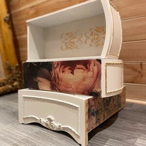 Puede incluir: Un cofre decorativo de tres niveles con acabado blanco y detalles dorados. El caj&oacute;n central presenta un gran estampado floral. El caj&oacute;n superior tiene un dise&ntilde;o floral dorado. El cofre est&aacute; sobre una superficie gris.