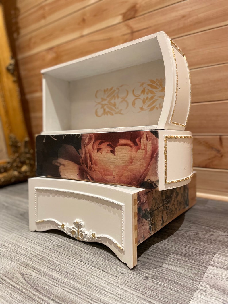 Puede incluir: Un cofre decorativo de tres niveles con acabado blanco y detalles dorados. El caj&oacute;n central presenta un gran estampado floral. El caj&oacute;n superior tiene un dise&ntilde;o floral dorado. El cofre est&aacute; sobre una superficie gris.