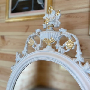 Puede incluir: Espejo ovalado con marco blanco y detalles ornamentados. La parte superior del espejo presenta un dise&ntilde;o de urna decorativa con detalles dorados. El marco tiene un tono rosa p&aacute;lido y el espejo est&aacute; sobre un fondo de madera.
