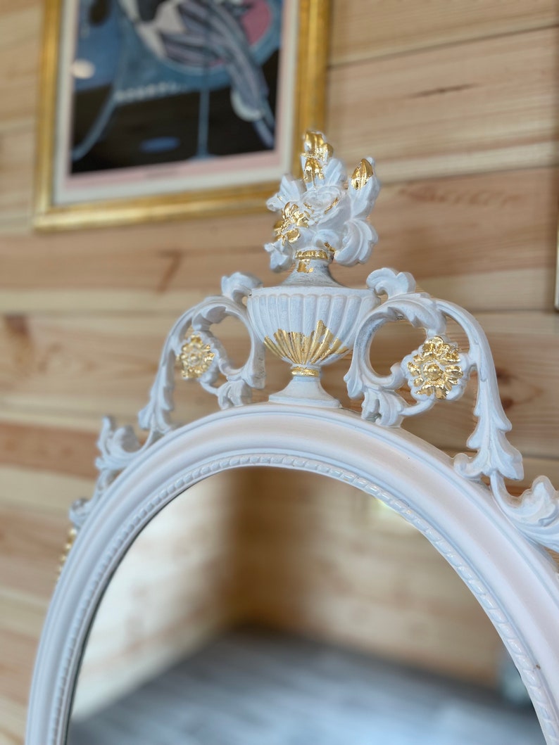 Puede incluir: Espejo ovalado con marco blanco y detalles ornamentados. La parte superior del espejo presenta un dise&ntilde;o de urna decorativa con detalles dorados. El marco tiene un tono rosa p&aacute;lido y el espejo est&aacute; sobre un fondo de madera.