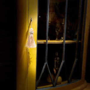 Pode incluir: Um arm&aacute;rio de canto amarelo com portas de vidro e moldura preta. Um pequeno tassel pendura em um gancho no lado do arm&aacute;rio.