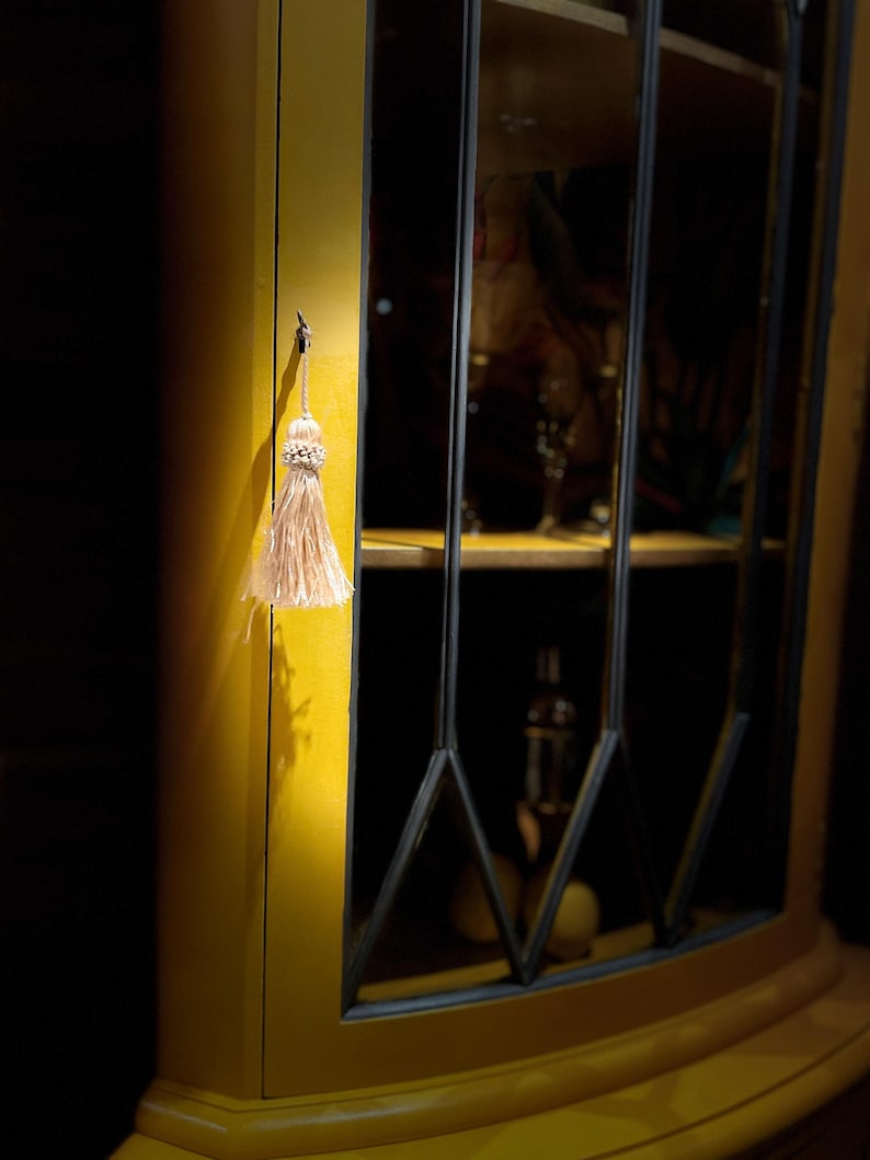 Pode incluir: Um arm&aacute;rio de canto amarelo com portas de vidro e moldura preta. Um pequeno tassel pendura em um gancho no lado do arm&aacute;rio.
