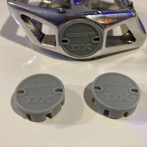 Puede incluir: Dos tapas de pedal de bicicleta Shimano DX de plástico gris. Las tapas tienen un logotipo Shimano DX en relieve.
