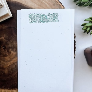 May include: A white sheet of paper with a green dragon design at the top. The paper has a speckled texture and is sitting on a wooden surface.