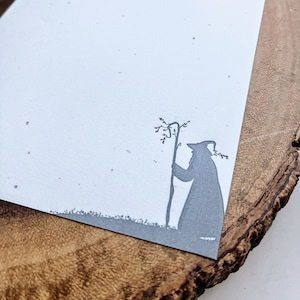 May include: A white card with a gray silhouette of a person holding a staff and standing next to a tree. The card is on a wooden surface.