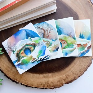 May include: Four square ceramic coasters with watercolor illustrations of hobbit homes in a winter landscape. Each coaster features a different scene with snow, trees, and a blue door. The coasters are displayed on a wooden surface.