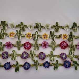 May include: Five horizontal strips of decorative trim featuring small, fabric-folded flowers in white, yellow, magenta, purple, and blue. Each strip is connected by green, textured, woven ribbon segments with small, vertical leaf-like accents.