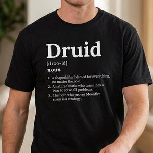 以下が含まれることがあります： 白字で「Druid」と定義が書かれた黒いTシャツ。定義には「shapeshifter」と「Moonfire spam」という言葉が含まれています。Tシャツは人が着ています。