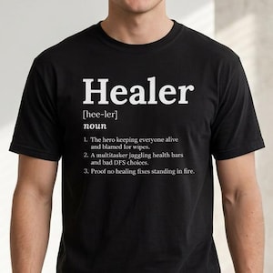 以下が含まれることがあります： 白字で「Healer」と定義が書かれた黒いTシャツ。定義には「hee-ler」という用語と、ヒーラーの役割を説明する3つの箇条書きが含まれています。画像に写っている人物はブルージーンズを着用しています。