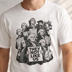 Könnte beinhalten: Weißes T-Shirt mit einer Schwarz-Weiß-Grafik historischer Figuren, die Schilder halten. Auf dem zentralen Schild steht „THEY NOT LIKE US“. Das Design umfasst Porträts von Männern in historischer Kleidung, mit einem Pferd und einer Kutsche im Hintergrund.