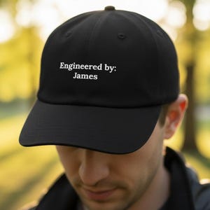 Puede incluir: Gorra de béisbol negra con visera curva. La gorra tiene el texto "Engineered by: James" en blanco, centrado en la parte delantera. El sombrero está hecho de un material negro liso.