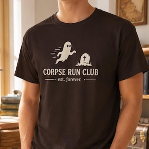 May include: A dark brown t-shirt featuring the words "CORPSE RUN CLUB" and a graphic of a ghost and tombstone. The text "est. forever." is below the main text. The shirt is worn with blue jeans.
