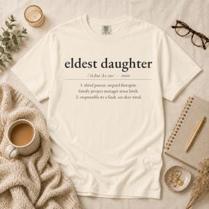 May include: A cream-colored short-sleeve t-shirt featuring the black text 'eldest daughter' followed by a dictionary-style definition including 'third parent, unpaid therapist, family project manager since birth' and 'responsible to a fault'.