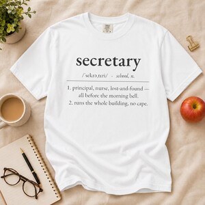 Op de afbeelding: Een wit t-shirt met korte mouwen en zwarte tekst die het woord secretary definieert. De tekst bevat een fonetische uitspraak en twee genummerde punten die de rol van het beheren van een schoolgebouw beschrijven, inclusief taken zoals het afhandelen van de directeur, verpleegkundige en gevonden voorwerpen.