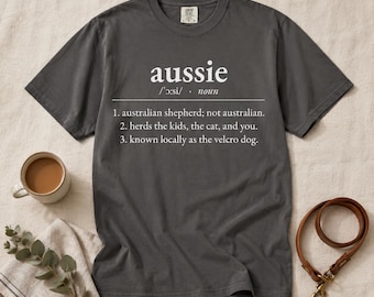 以下が含まれることがあります： チャコールグレーの半袖Tシャツ。白い文字で「aussie」をオーストラリアン・シェパードと定義し、子供や猫や人を追いかけ、ベルクロドッグとして知られていると記されている。クルーネックで標準的なフィット感のシャツ。