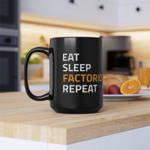 Op de afbeelding: Zwarte koffiemok met witte tekst die "Eat Sleep Factorio Repeat" luidt. De tekst "Factorio" is oranje.
