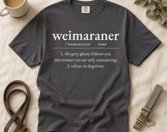 以下が含まれることがあります： チャコールグレーの半袖Tシャツ。白い文字で「weimaraner」という単語が名詞として定義されており、発音記号と、この犬種に関する2つの説明文（グレーゴーストやベルクロについての言及）が記載されています。