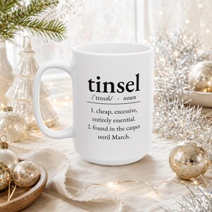 May include: A white ceramic mug featuring black printed text that defines the word tinsel as cheap, excessive, and entirely essential, noting it is found in the carpet until March. The mug has a standard handle and a smooth, cylindrical body.