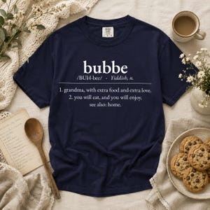May include: A navy blue short-sleeve t-shirt featuring white text that defines the word bubbe as a grandma with extra food and extra love, noting you will eat and enjoy, with a reference to home. The shirt has a crew neckline and a label.